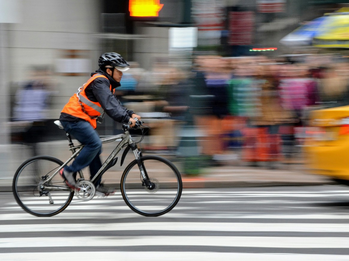 Ein Radfahrer mit Warnweste und Helm fährt über einen Zebrastreifen hinter einem gelben Auto.