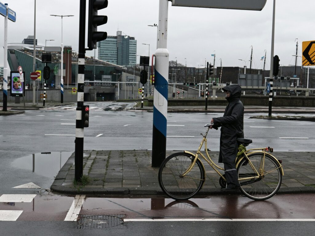 Eine Straßenkreuzung im Regen. Eine Radfahrerin mit Regenkleidung steht an der Ampel und wartet.